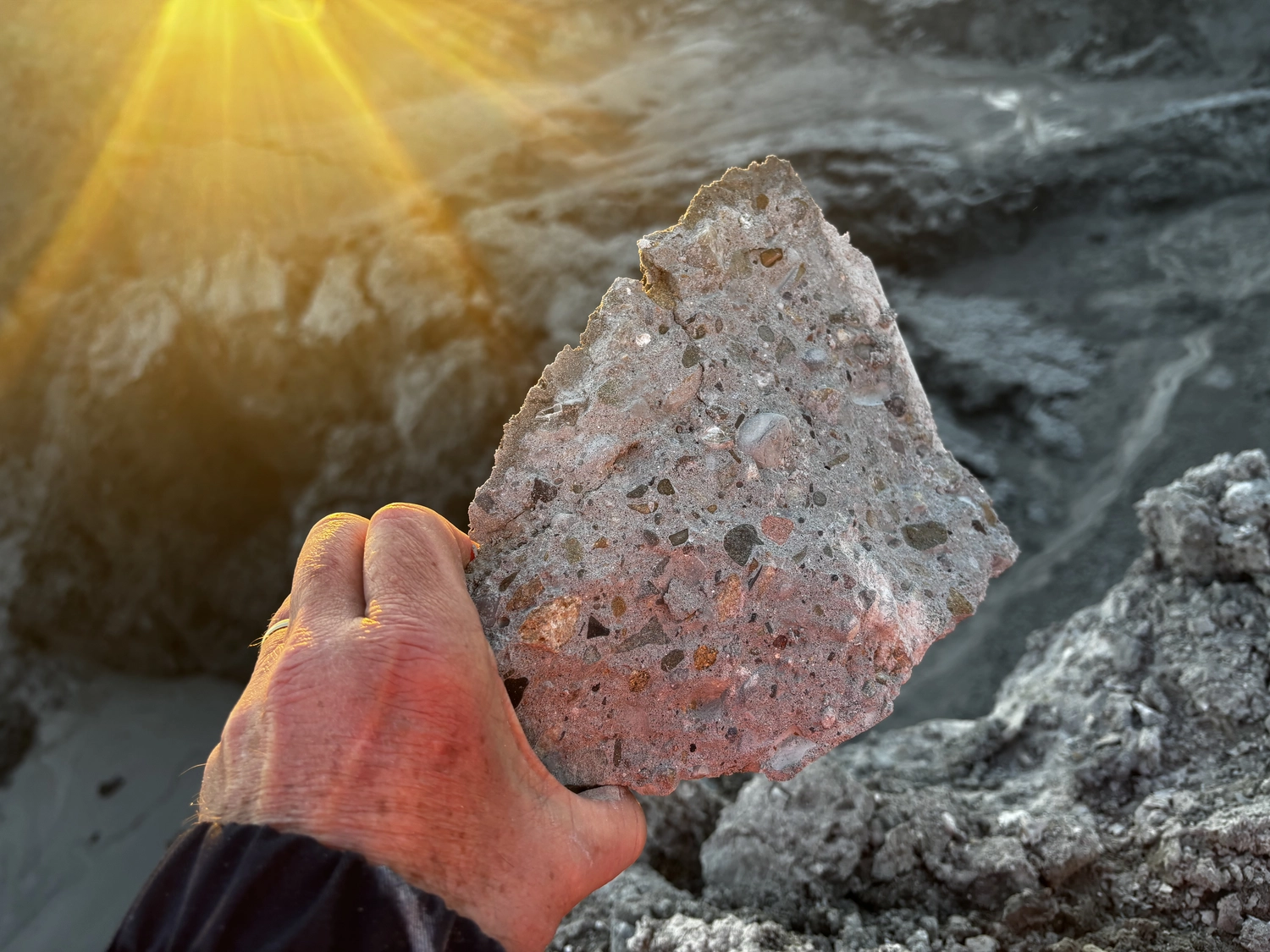 Dr. Rehm – Tunnelbau-Service – Eine Hand hält ein großes, grobes Stück Beton- oder Steinbruch vor einem rauen, sonnenbeschienenen Hintergrund hoch.