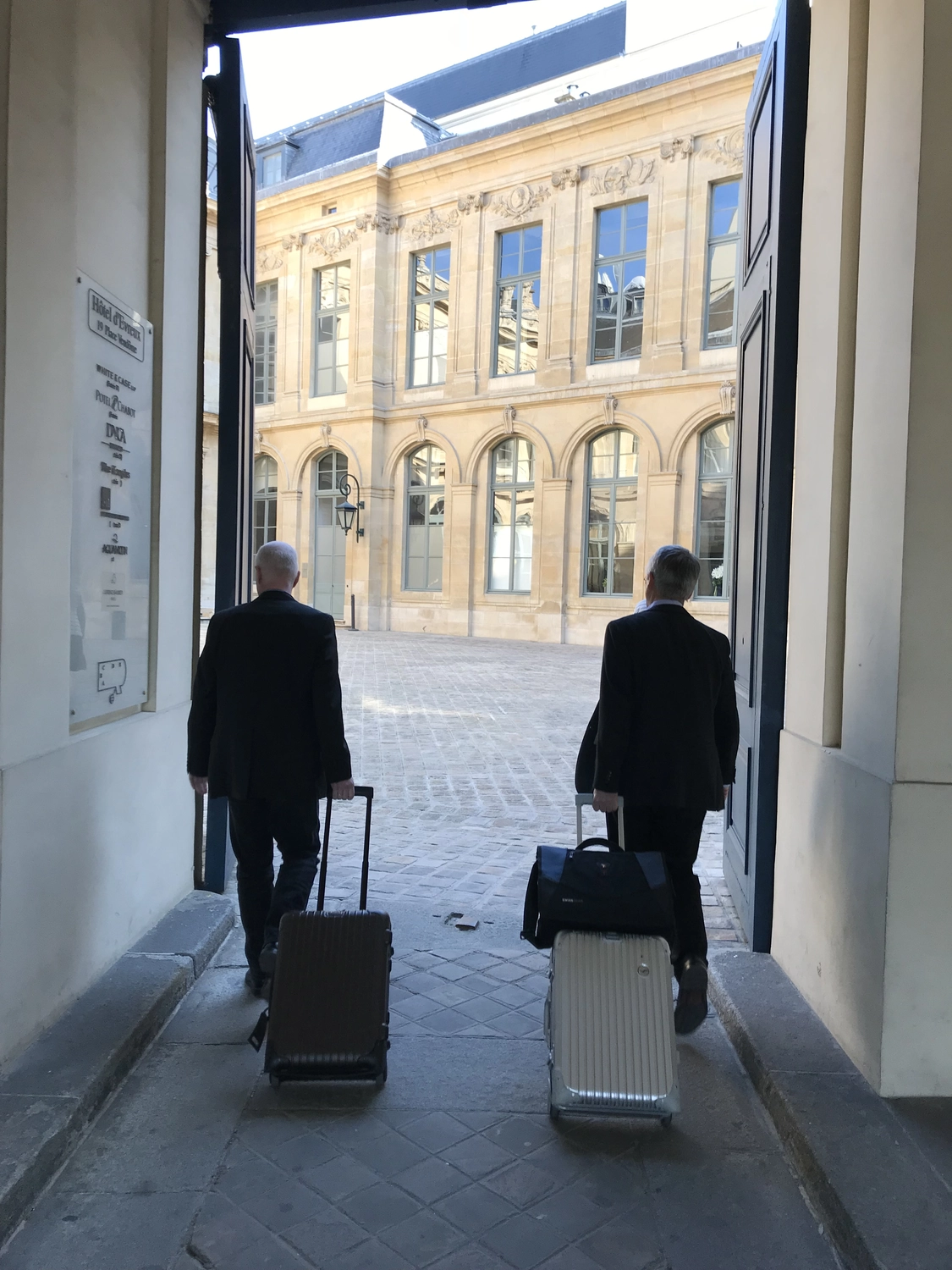 Dr. Rehm – Tunnelbau-Service – Zwei Männer in Anzügen rollen Gepäck aus einem Gebäudeeingang in einen gepflasterten Innenhof.